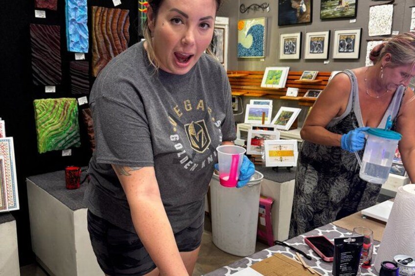 A person in a grey t-shirt with a logo, holding a pink cup, and another person in a patterned dress with gloves, working at a table with various items. 4