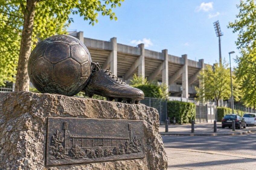 A soccer ball and cleats on a stone pedestal with a plaque, set against a backdrop of a stadium and urban street. 3