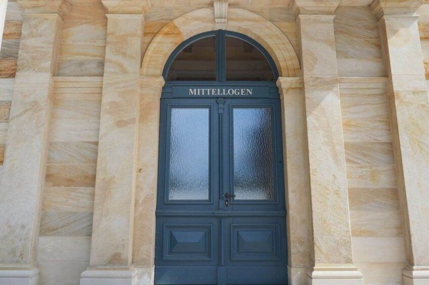 A dark blue door with the inscription 'MITTELLOGEN' above it, set within a classical architectural style with marble columns. 5