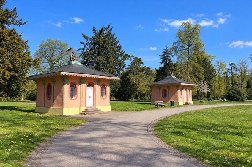 Two small, pinkish buildings with arched windows and a central door, surrounded by a grassy park with a paved path leading to them. 2