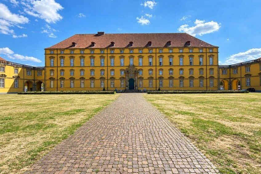A large yellow building with a central entrance, surrounded by a paved courtyard and grassy areas. 5