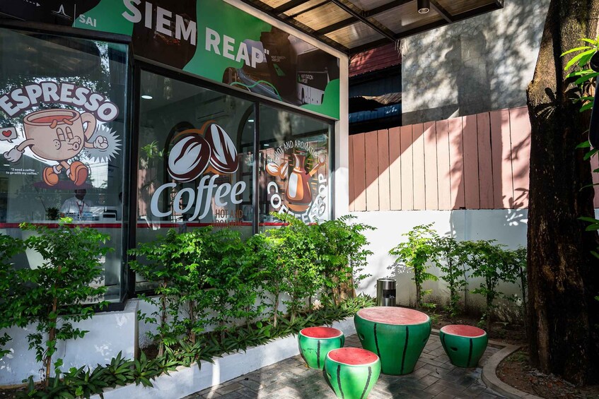 A coffee shop with a large window displaying coffee-related artwork and the name 'SIEM REAP'. There are potted plants and a small outdoor seating area with round tables. 4