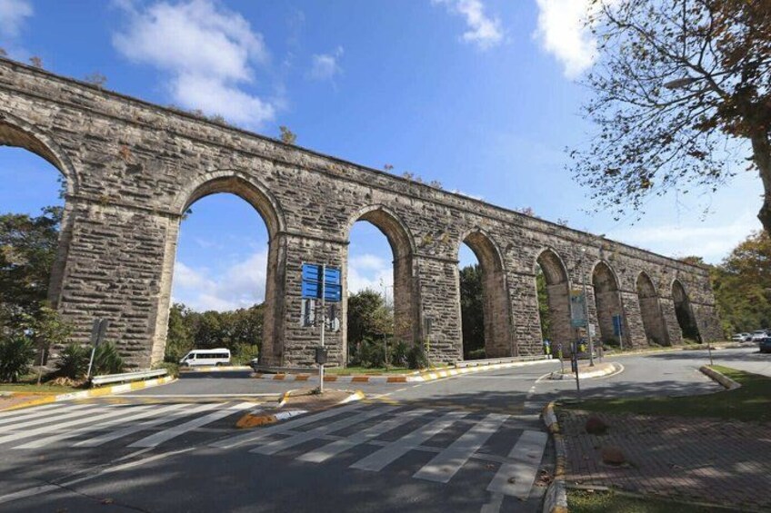 A stone aqueduct with multiple arches, a road with a pedestrian crossing, and a white van in the foreground. 3