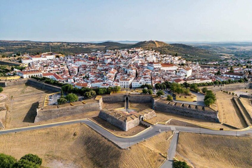 A town with a prominent fortification, surrounded by a mix of white and red buildings, and a hilly landscape in the background. 1