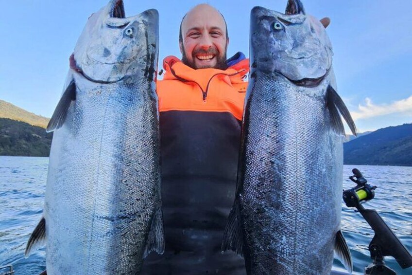 A person holding two large fish on a boat with a lake and mountains in the background. 5