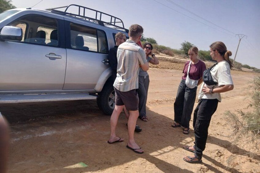 A group of people standing around a silver SUV on a dirt road in a desert-like environment. 2
