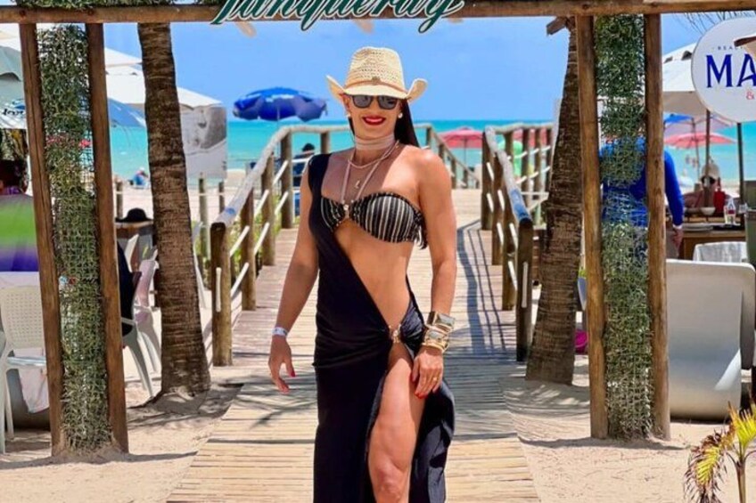 A person in a bikini and hat on a wooden boardwalk with palm trees and beach umbrellas in the background. 2