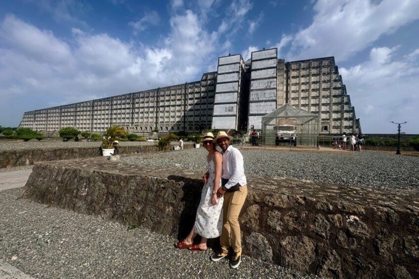 A large, rectangular building with a grid-like facade, a smaller structure with a glass roof, and a rocky terrain with a few people standing. 1