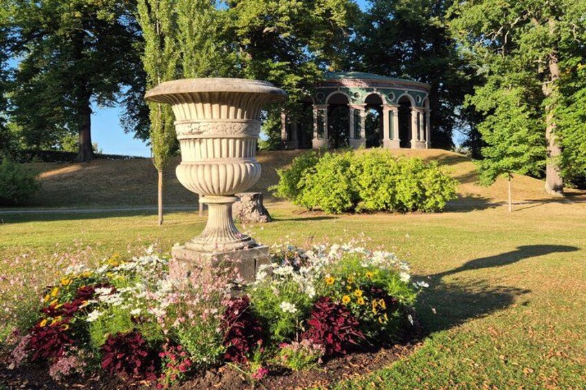 A garden with a large urn, colorful flowers, and a gazebo in the background. 1