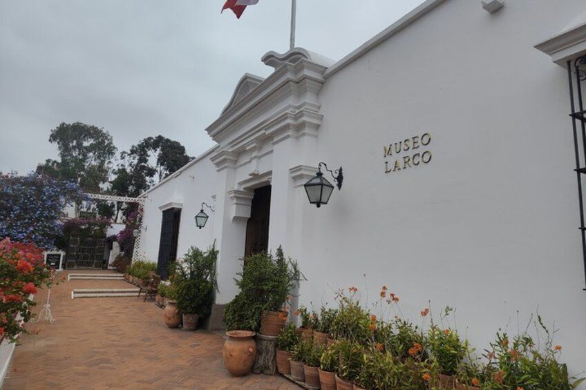 A white building with the sign 'MUSEO LARCO' on its facade, surrounded by potted plants and a brick pathway. 1