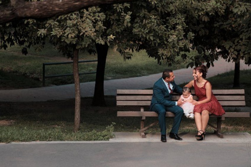 A family of three sitting on a bench under a tree in a park. 4