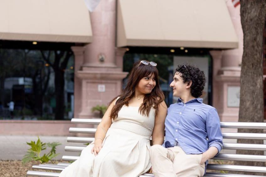Two people sitting on a bench with a building in the background. 5