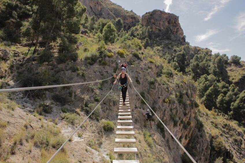 A person walking on a rope bridge over a canyon with rocky cliffs and sparse vegetation. 1