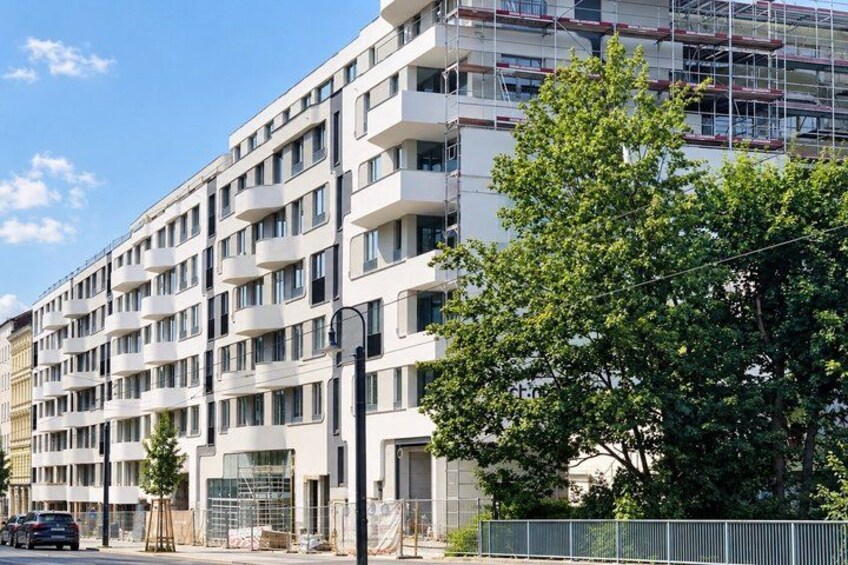 A multi-story building with balconies, a tree in the foreground, and a construction site to the right. 3