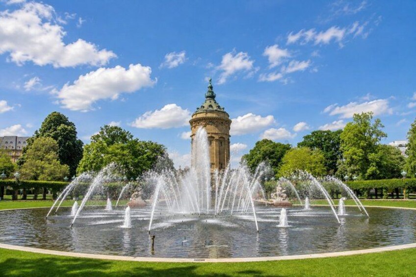 A fountain with water jets in a circular basin, surrounded by a park with trees and a building in the background. 4