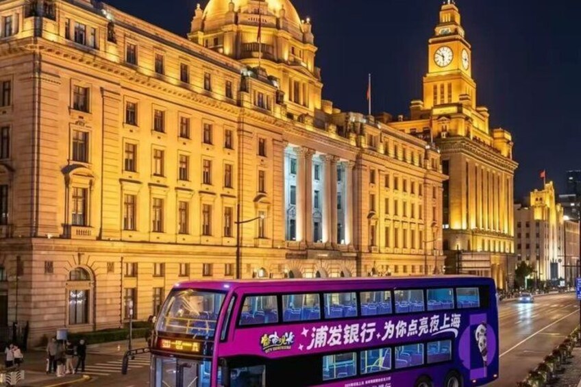 A purple double-decker bus in front of a large, illuminated building with a clock tower. 1
