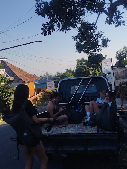 A group of people sitting on a vehicle's bed in a rural setting with a signboard in the background. 3