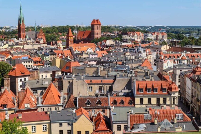 A densely packed cityscape with red-roofed buildings, a prominent church spire, and a bridge in the background. 2