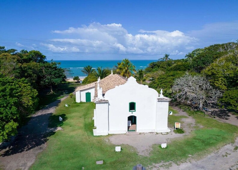 A white church with a tiled roof surrounded by lush greenery and a view of the ocean. 4