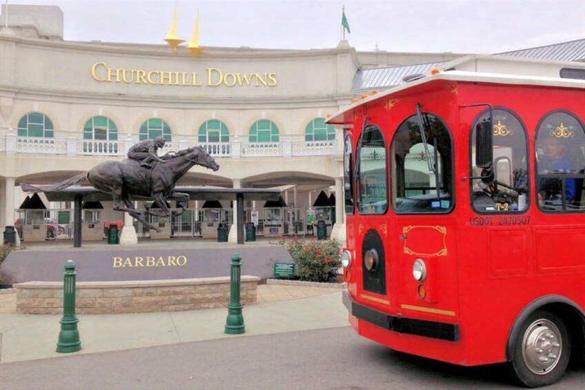 A red vintage bus in front of Churchill Downs, with a statue of a jockey and horse, and the name 'BARBARA' on a sign. 1