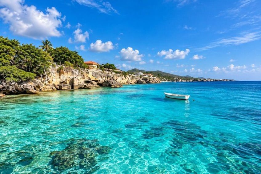 A clear turquoise sea, a small boat, rocky cliffs, and a distant building. 1