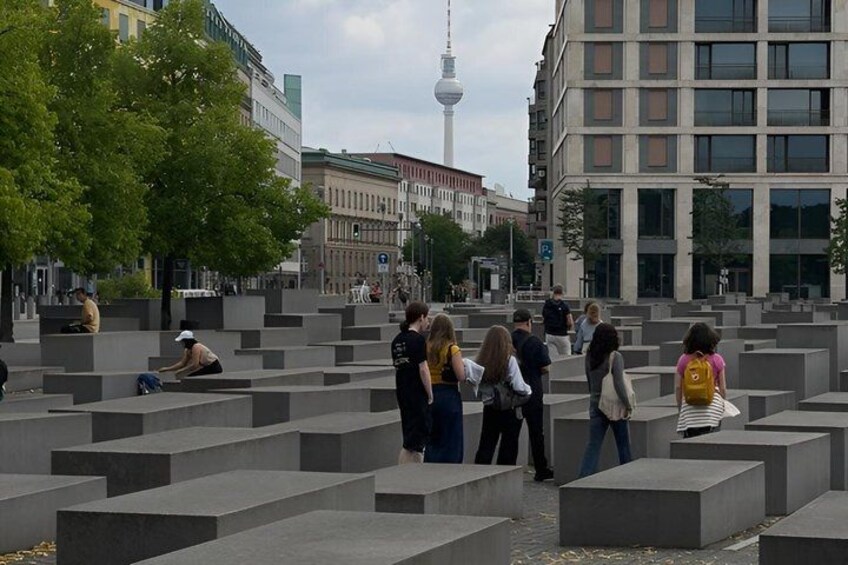 A public square with people sitting and walking, a modern building, and a tower in the background. 2