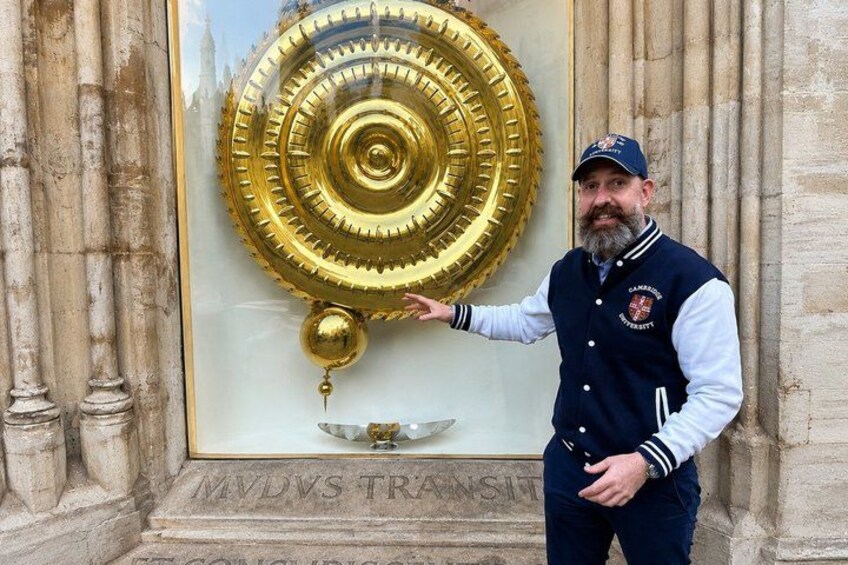 A man standing next to a large golden object with a circular design, in front of a stone wall with inscriptions. 1