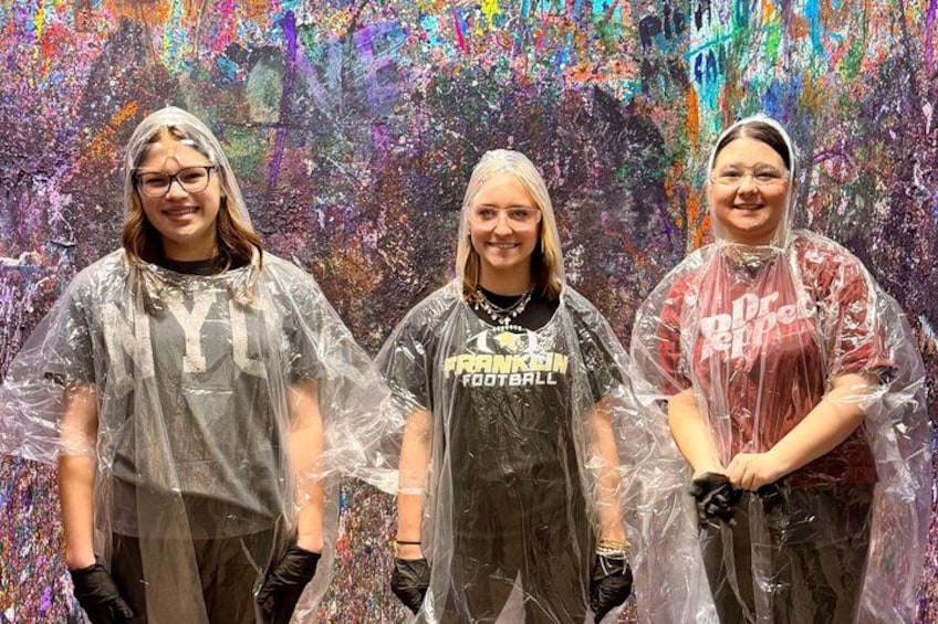Three people wearing t-shirts and rain ponchos in front of a colorful abstract painting. 4