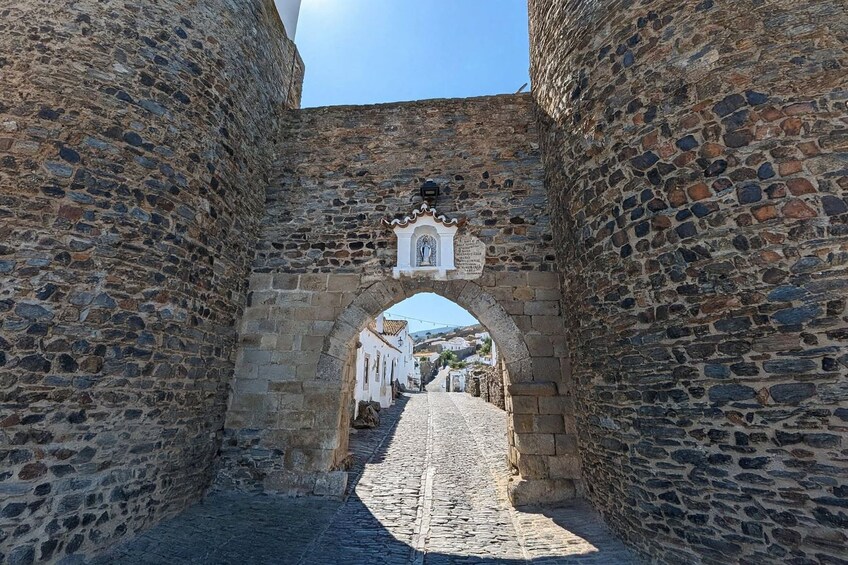 A stone archway leading to a cobblestone street with buildings in the background. 1