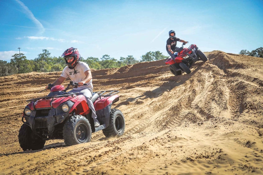 Two people riding red quad bikes on a dirt track with a clear sky. 1