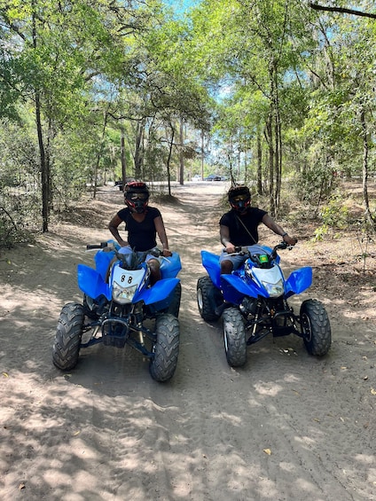 Two riders on blue ATVs on a dirt path surrounded by trees. 3