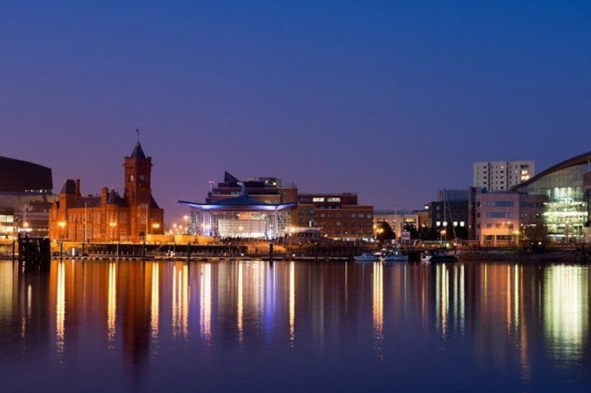 A cityscape at dusk with a prominent historic building, a modern glass structure, and reflections on the water. 5