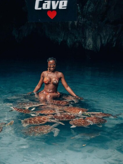 Een persoon in een bikini, omringd door zeeschildpadden in een zwembad, met een bordje met de tekst 'grot' op de achtergrond. 1
