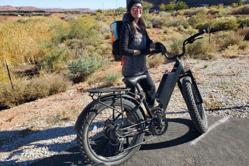 A person standing next to a black electric bicycle with a rear rack, in a desert-like environment with sparse vegetation and a clear sky. 5