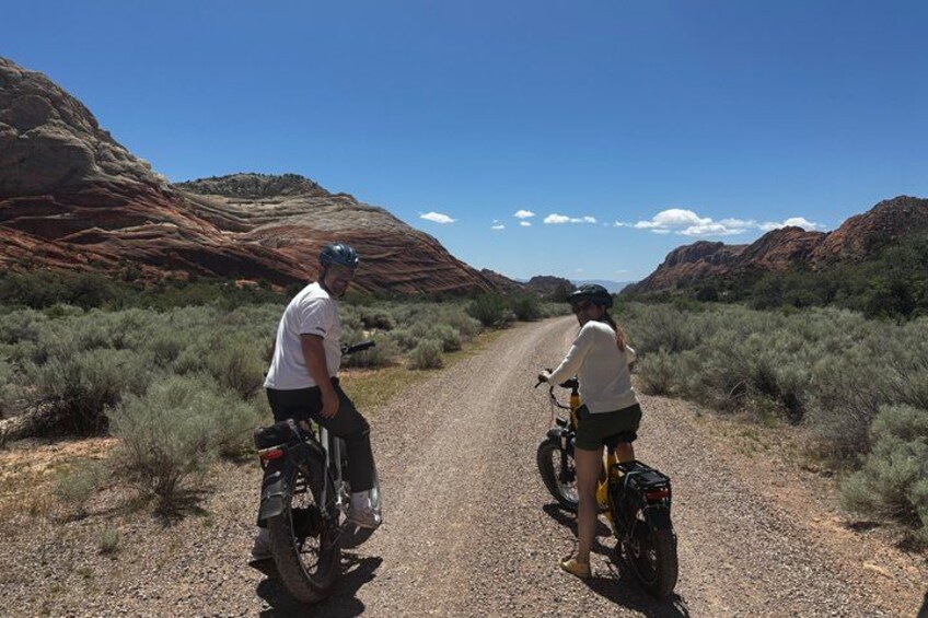 Two cyclists on a gravel road with red rock formations in the background. 4