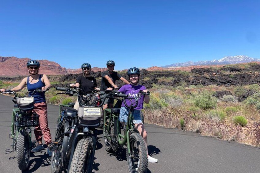 Four people on mountain bikes on a paved road with a desert landscape and mountains in the background. 2