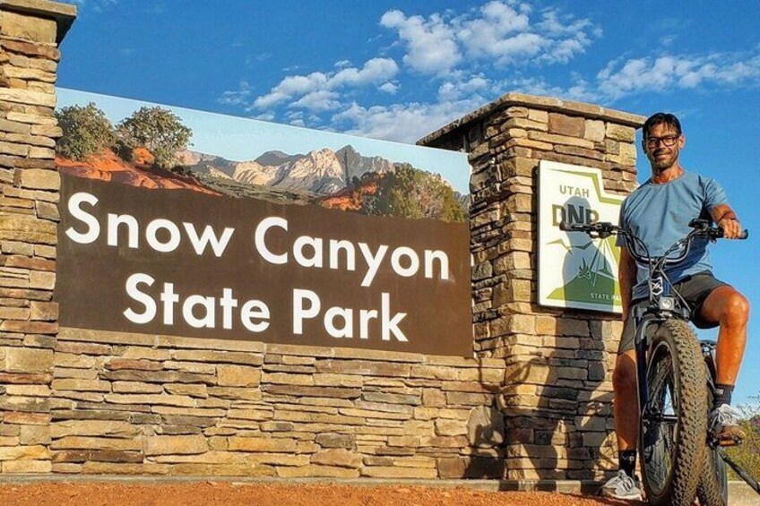 A person on a bicycle in front of a sign for Snow Canyon State Park. 1