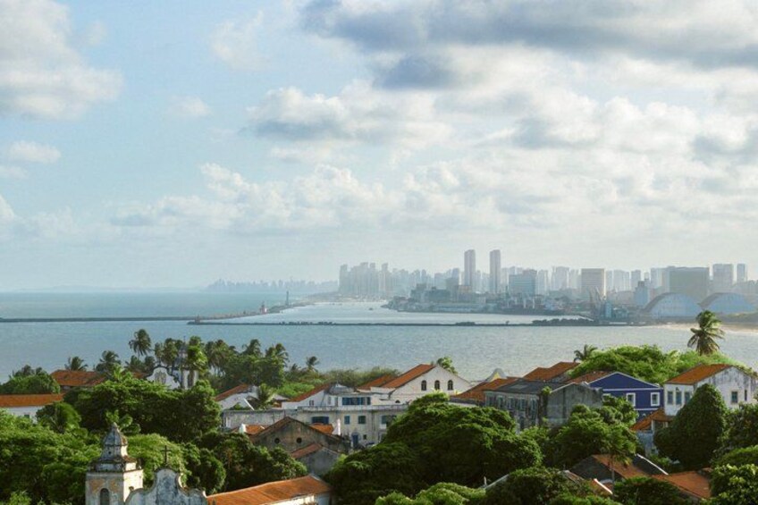 A coastal cityscape with historic buildings, a marina, and a distant urban skyline. 1