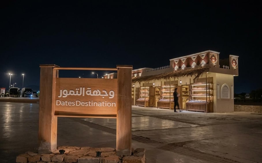 Puuinen kyltti, jossa on arabiankielistä tekstiä ja englanninkieliset sanat »Dates Destination», on sijoitettu koruliikkeen eteen yöllä. 2