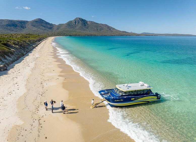 Ein Strand mit einem Boot namens 'Southern Cross' an der Küste, Menschen, die am Strand entlanggehen, und Bergen im Hintergrund. 1