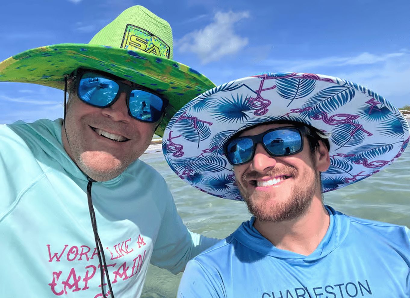 Two people wearing hats and sunglasses, one in a light blue shirt and the other in a green shirt, standing near water. 3