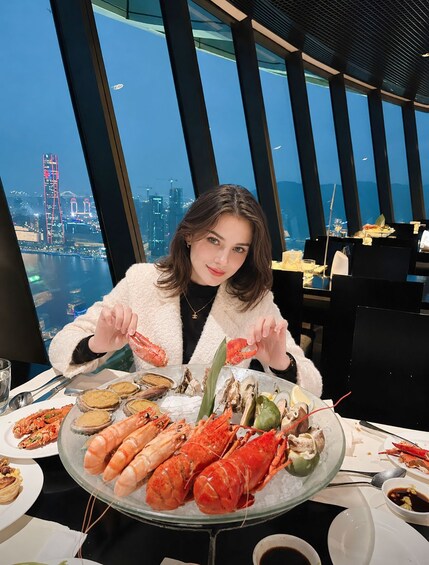 A person dining at a table with a seafood platter, a cityscape view, and a modern building with a distinctive red tower in the background. 1