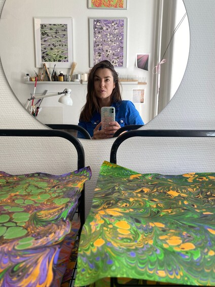 Une personne prend un autoportrait dans une pièce où l’on aperçoit un miroir, un bureau recouvert d’une nappe colorée et diverses fournitures artistiques rangées sur des étagères. 1