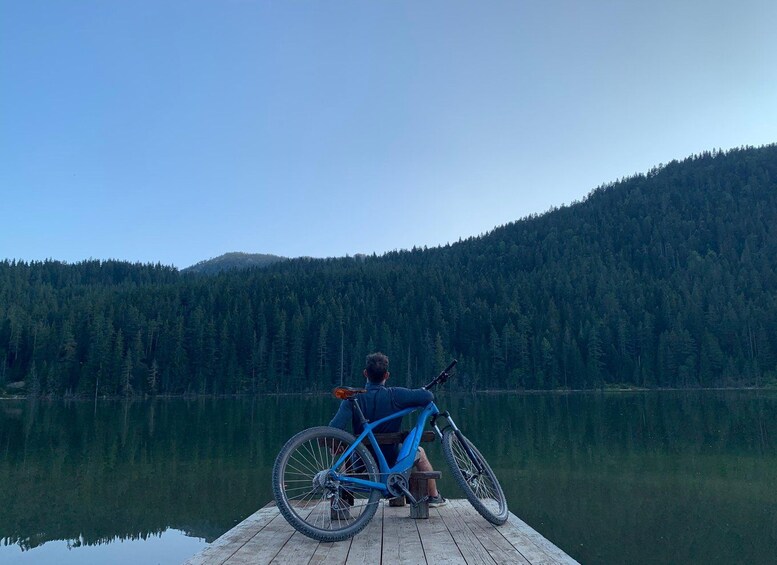 Una persona sentada en un muelle con una bicicleta, mirando un lago tranquilo y montañas boscosas. 3