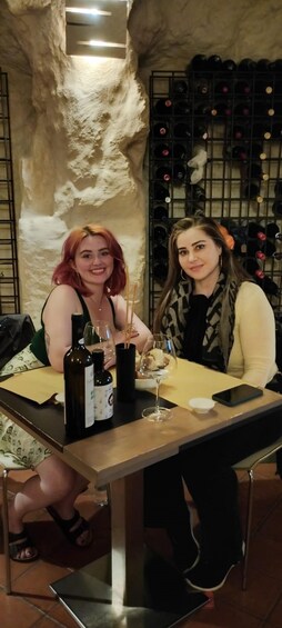 Two people sitting at a table with wine bottles and glasses. 1