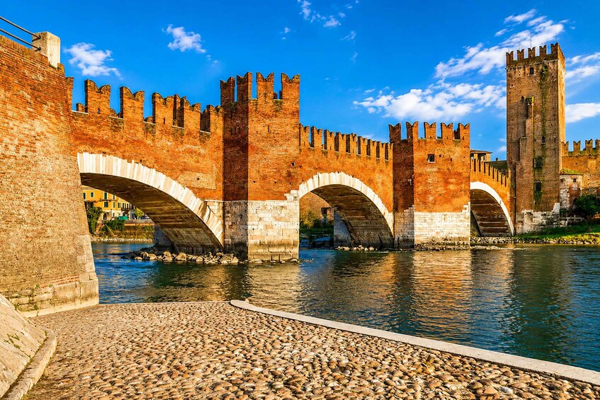 Un puente de piedra histórico con arcos sobre un río, flanqueado por muros de ladrillo y una alta torre. 4