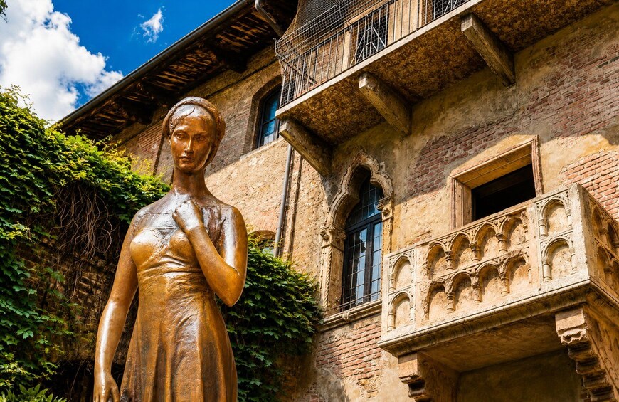 Una estatua de bronce de una mujer con vestido, un edificio de ladrillo con balcón y un cielo azul despejado. 1
