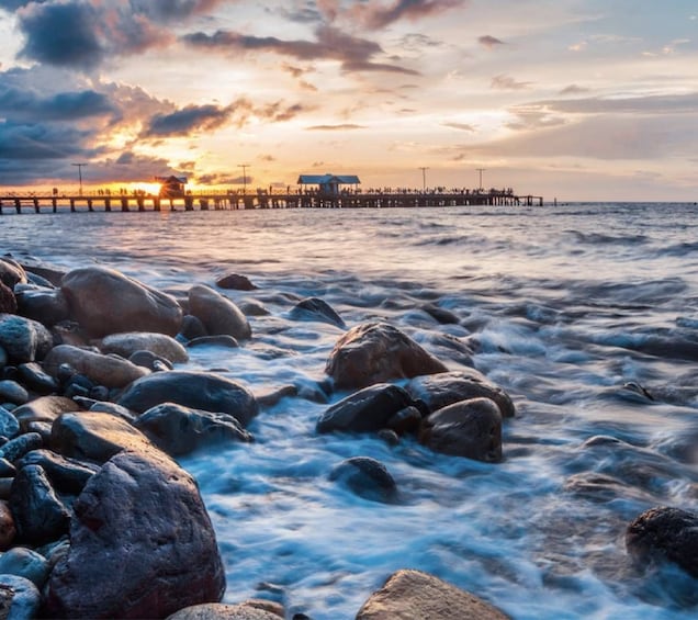 Een rotsachtige kustlijn met golven, een pier die het water in reikt, en een zonsondergang op de achtergrond. 3