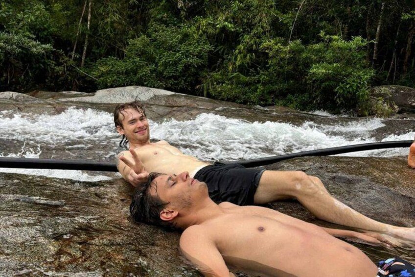 Two people relaxing on a rock by a flowing river in a forested area. 3
