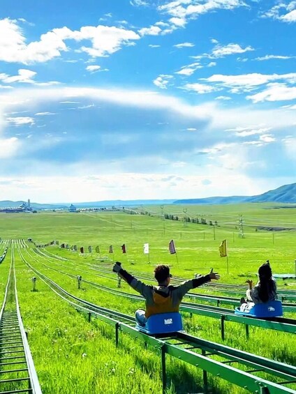 Two people sitting on a green metal bench with their arms outstretched, overlooking a railway track and a grassy field with flags. 3